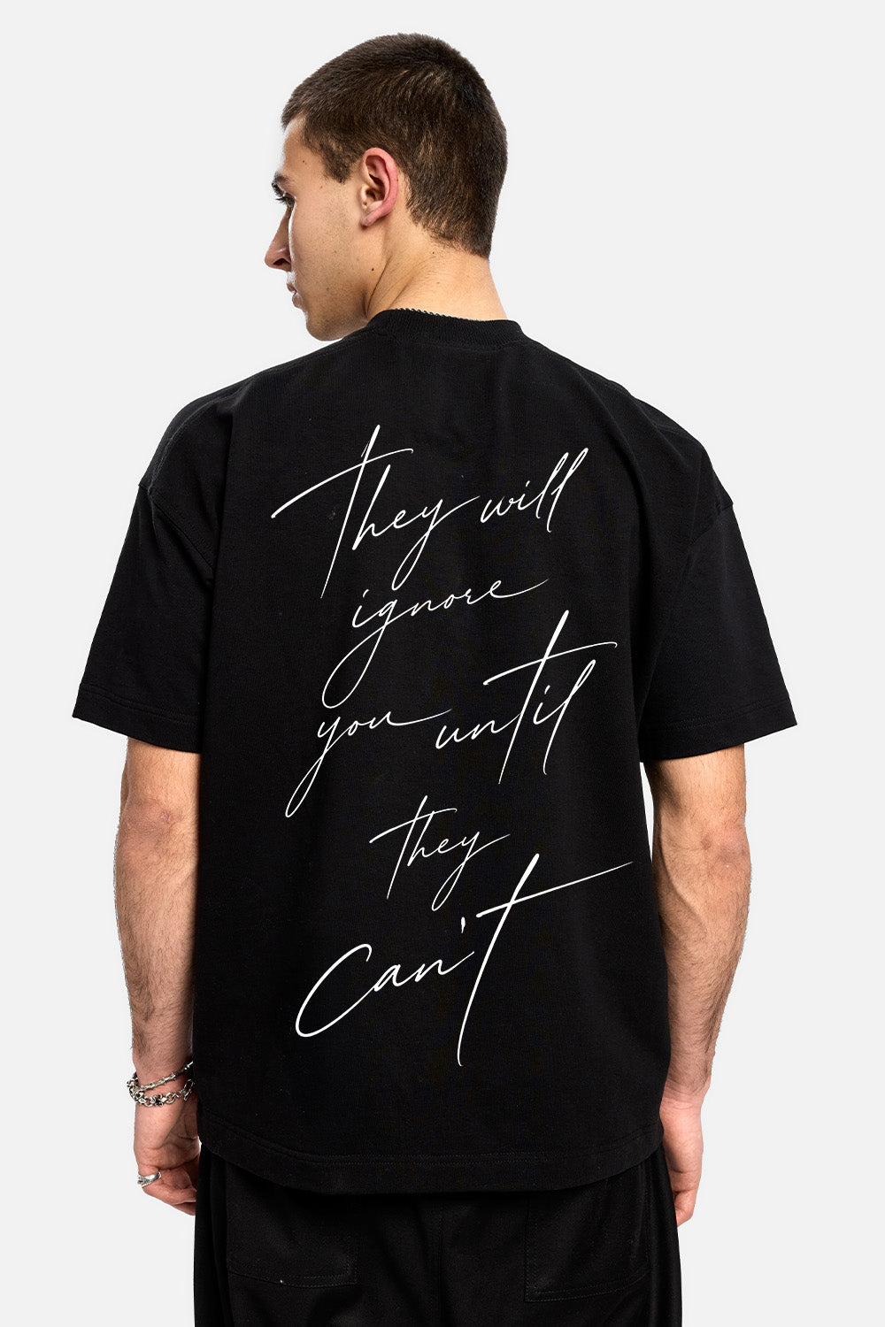 Vedere din spate a unui model purtând tricoul oversized negru They Will Ignore You pentru bărbați, evidențiind mesajul grafic scris de mână