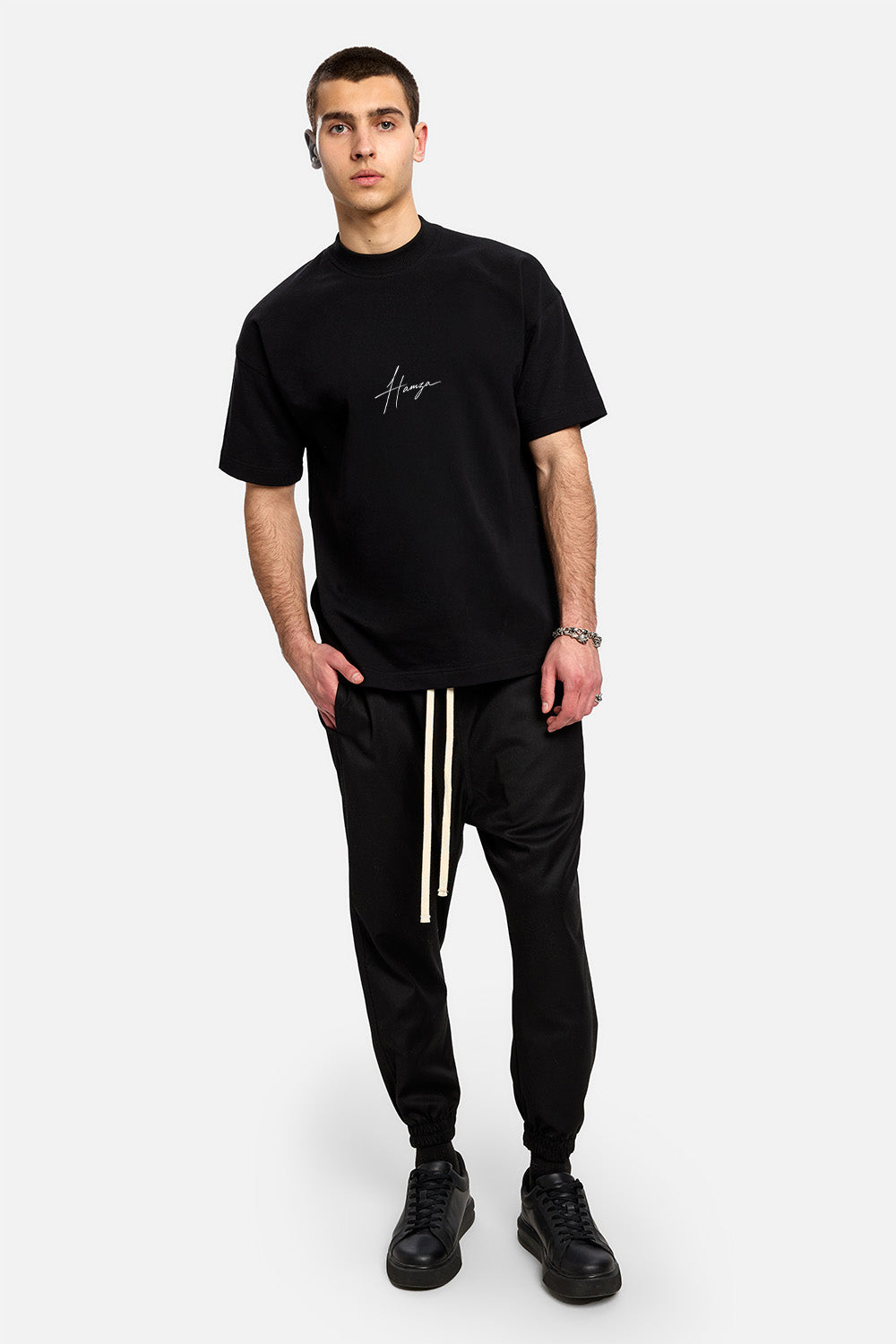 Vedere frontală full-body a unui model purtând tricoul oversized negru They Will Ignore You pentru bărbați, cu semnătura Hamza pe față